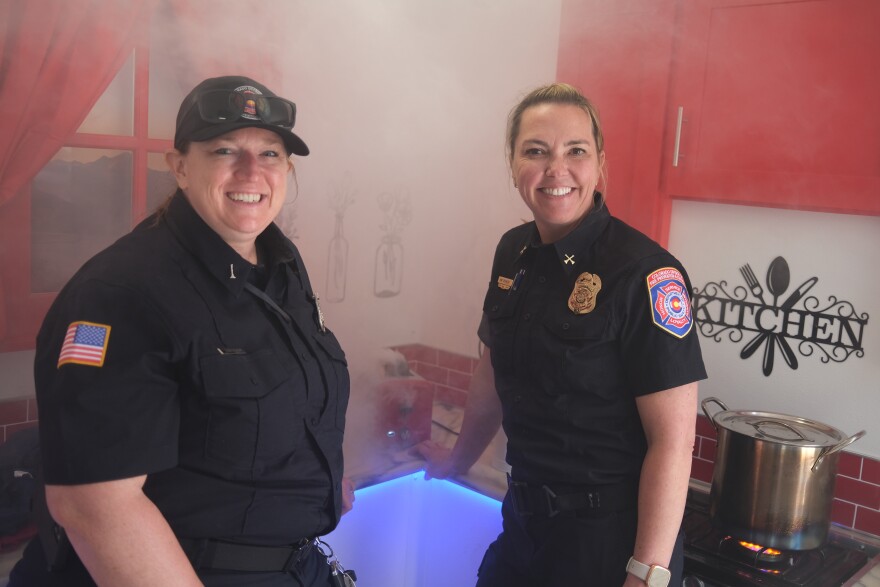 (L to R) Jen Rand and Kimberly Spuhler with the Colorado Division of Fire Prevention and Control standing in the Fire Safety Simulator’s mock kitchen on April 30, 2026. Behind them, a toaster emitting fake smoke offers hands-on practice for dealing with an electrical fire.