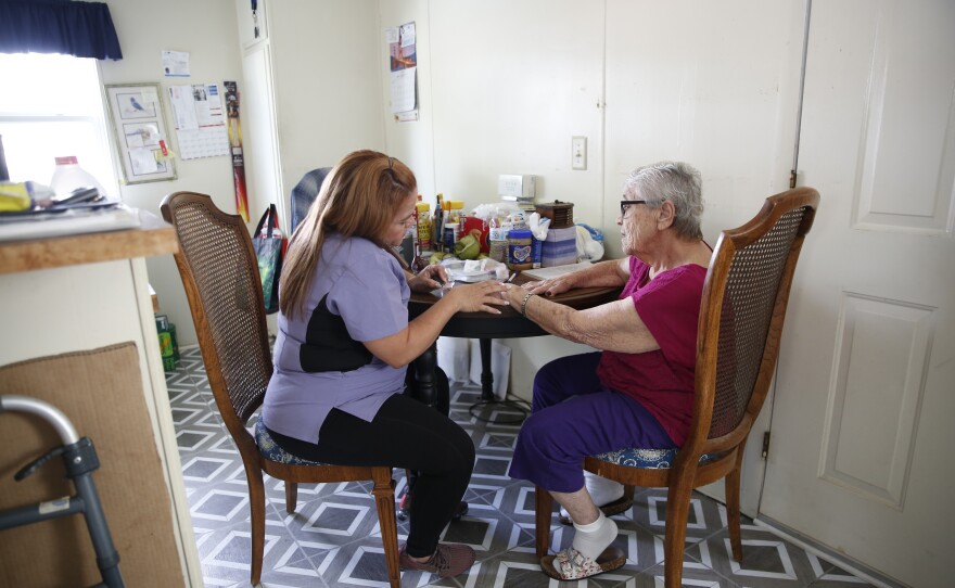 A woman helps take care of an elderly woman at a dining room table. 