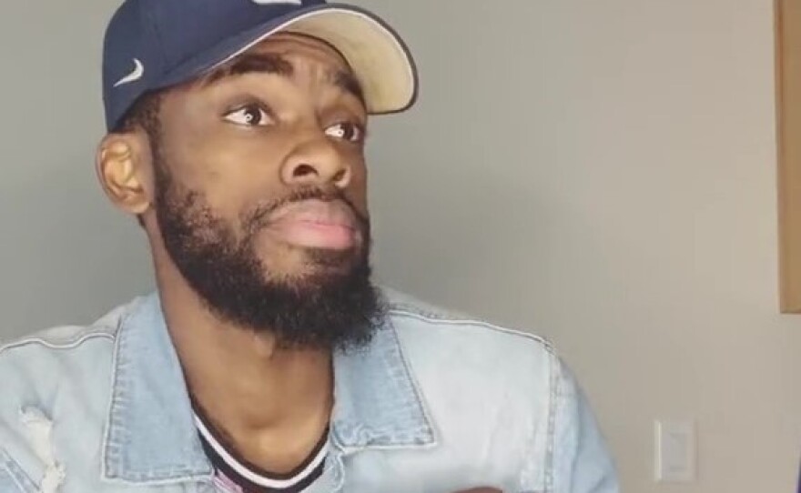 A young Black man looking past the camera. He has a jean jacket on and an arm across his chest.