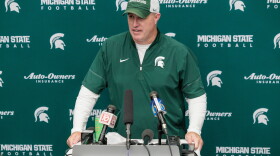 Pat Fitzgerald speaks at a podium with several microphones on it in front of a backdrop with MSU logos. He is wearing a MSU shirt and hat.