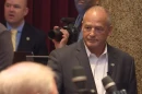 Gary Pinkel is seen in front of microphones. There is a photographer in the background with a man in a suit. Pinkel has a neutral expression.  
