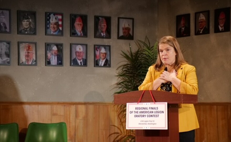 A woman wearing a yellow coat standing in front of a podium with pictures of military servicemen in the background.