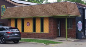 Photo of low-slung brick building with warm green and yellow wood trim, on a street with a parked grey car. WMUK has blurred the license plate number.