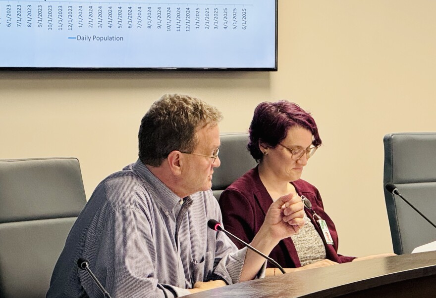 Two people sit at a conference table with microphones in front of them. A man speaks while gesturing, and a woman takes notes. A screen displaying a line graph is visible in the background.