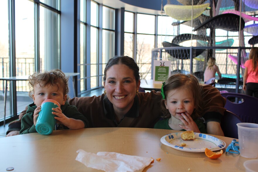 A woman kneels behind a table holding two sitting toddlers inside her arms while smiling. Food is on the table and the child on the left is drinking from a bottle. 