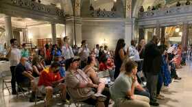 Dozens of people sit in folding chairs, and approximately half stand with their right hand held aloft. 