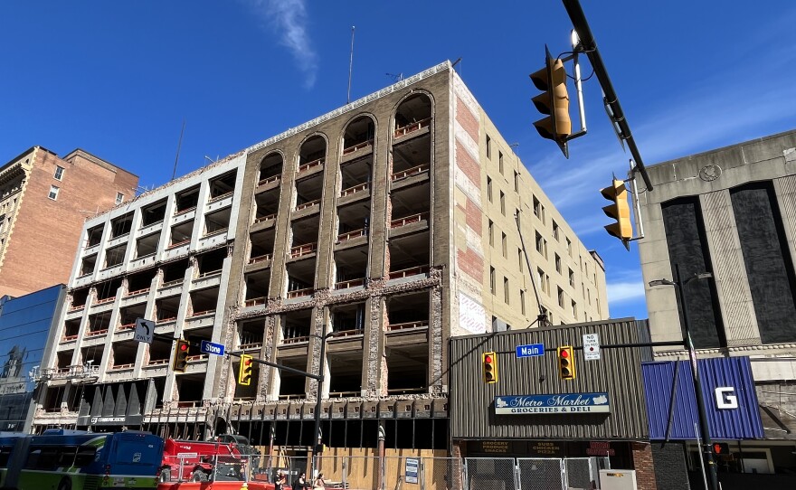 A photo of the section of East Main Street opposite Stone Street shows two buildings that used to be covered with glass panels now with century-old facades exposed. One building is a white, boxy grid. The other has arches windows cut at the top floor. Barricades and fencing surround the buildings at street level.