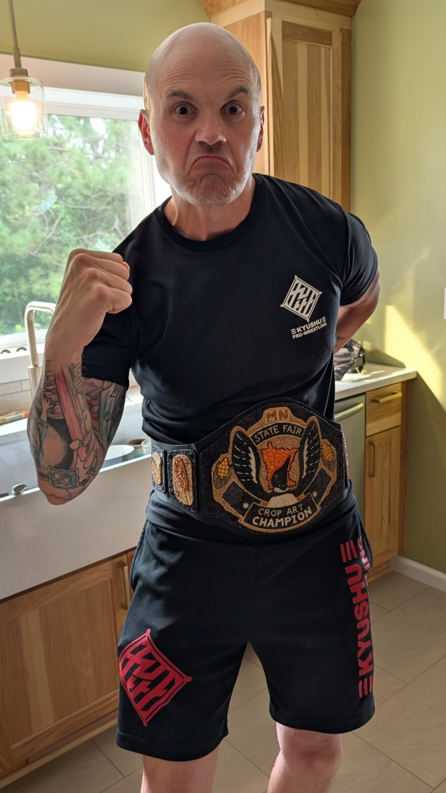 A man in black shorts and t-shirt pulls a tough face while wearing a wrestling championship-style belt adorned with a loon