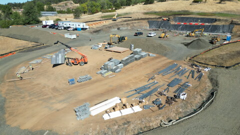 An overhead view of a dirt construction site is shown, with piles of materials as well as construction vehicles. 