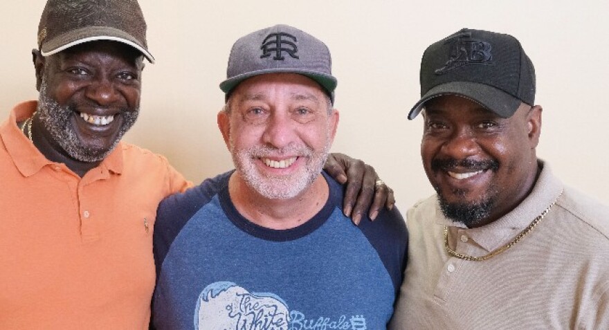 Three men standing side-by-side, wearing baseball caps and smiling into the camera