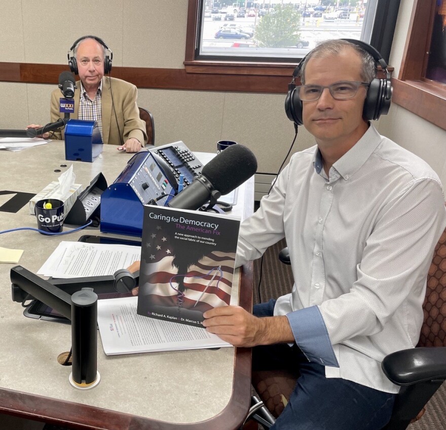 Two men wearing headphones sit at a table in a radio talk show studio: a man at left is wearing a tan blazer and a white, purple, and green plaid button-down shirt; a man at right has short dark hair and is wearing glasses, a white long-sleeved button-down shirt with light blue cuffs, polka dots, and jeans. He is holding a book with an American flag on the front cover. The book is titled "Caring for Democracy."