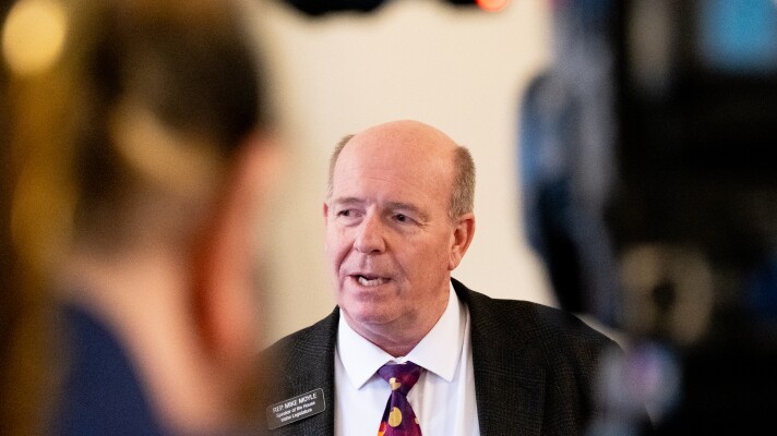 A balding man in a gray suit framed between a television camera and the camera operator.