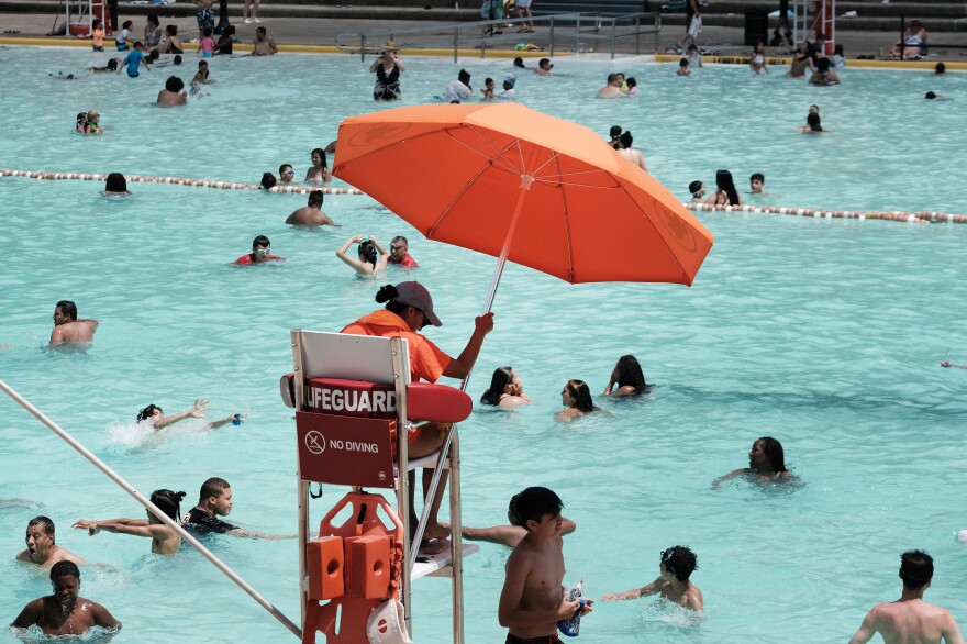 People swim in the light turquoise water of a public pool, as a lifeguard sits in a tall chair holding an orange umbrella.