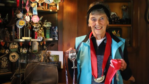 An elderly woman wearing workout gear and a medal is smiling and holding two medals. Behind her is a trophy case filled with awards.