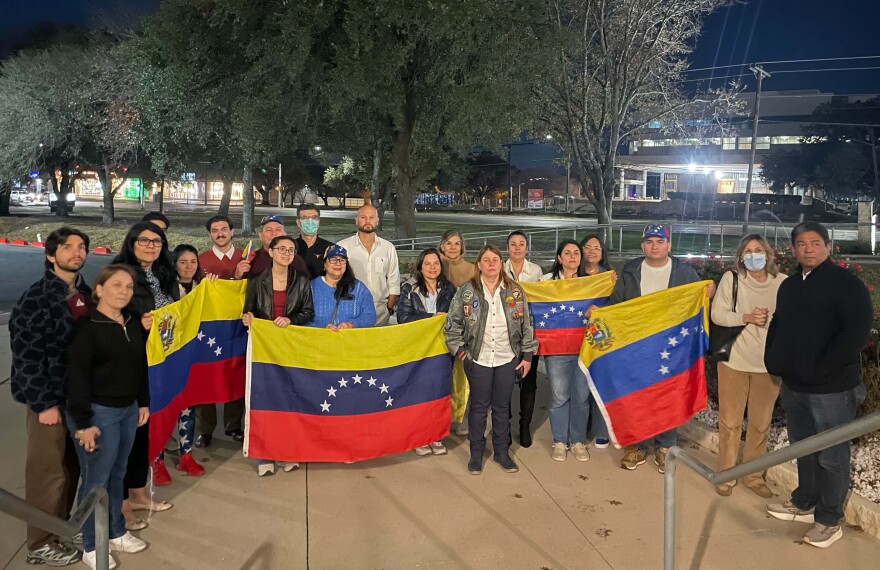 A group of 16 people stands outside at night, with several people holding large Venezuelan flags in yellow, blue and red.