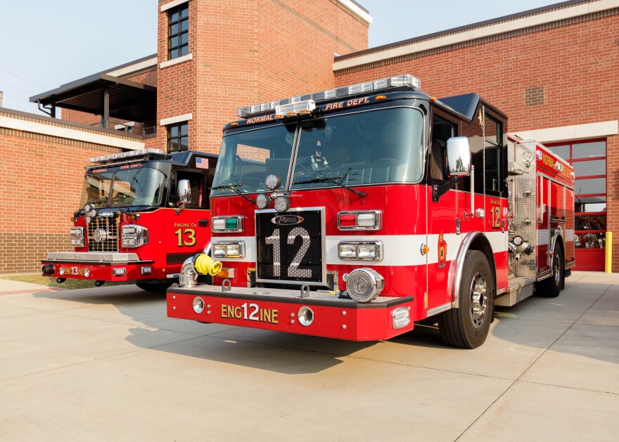 Two red fire trucks are parked in front of a brick firehouse. 