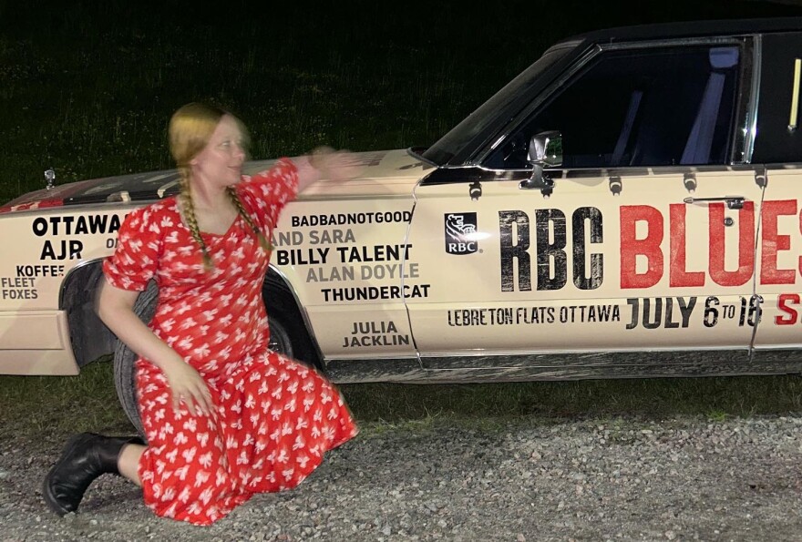 A woman in a print dress kneels next to an older sedan with musicians' names painted on the side.