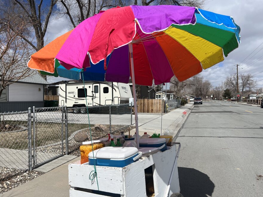 A street food cart with a multi colored umbrella sitting on a residential street with driveways in the background.