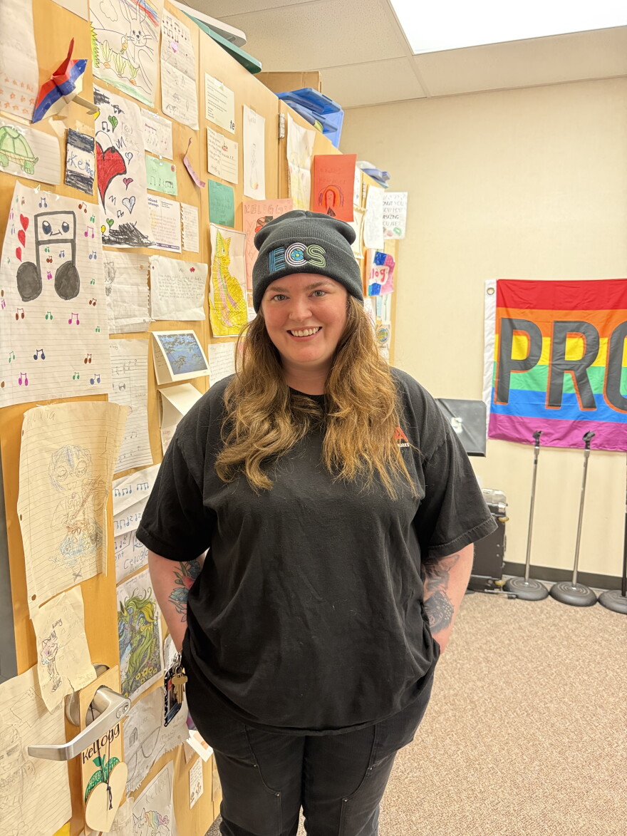 A person stands smiling with their hands in their pockets, in front of a wall with children's art posted on it. 