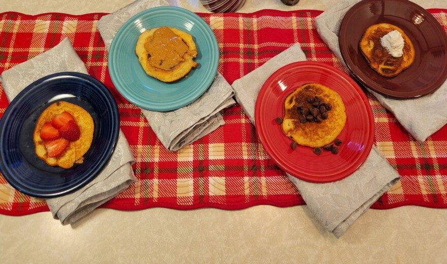  Four plated pancakes, each with a different topping (strawberries, peanut butter, chocolate chips, whipped cream)