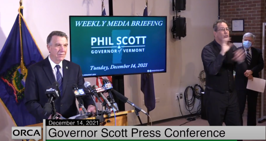 A man stands delivering a press briefing at a podium while another man signs his comments.