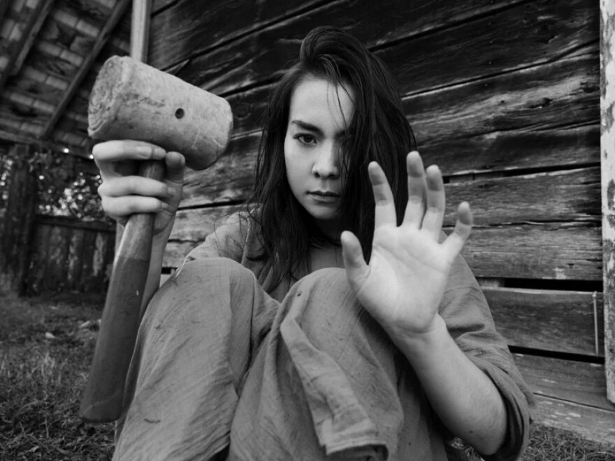 A woman sitting on the ground near the outside wall of a wooden structure holds a wooden mallet in one hand with her other hand raised in surrender.