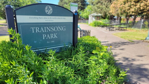 A sign for Trainsong park, along with bushes, sidewalk, picnic tables in the background.