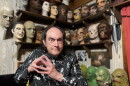 A man in a skeleton-patterned shirt poses in front of a wall of Halloween masks.