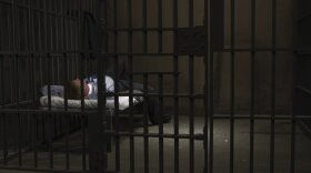 A man lies down in a prison cell with one leg propped up and one foot on the floor. 