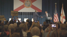 Florida Gov. Ron DeSantis shakes hands with U.S. Rep. Byron Donalds onstage in front of a large Florida flag as audience members watch and take photos.