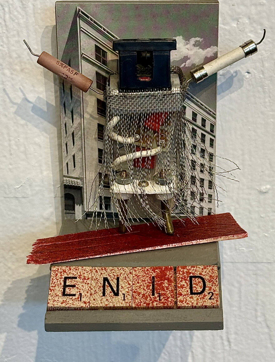 A tiny electronic figure made of wire mesh, ceramic fuses, and resistors stands atop vintage Scrabble tiles spelling "ENID," set against a cityscape photograph.