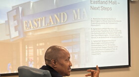 In the foreground, a man addresses someone offscreen. A slideshow is displayed behind him 