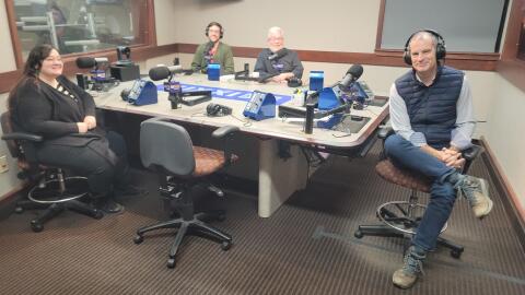 Four people sit at a table in a radio talk studio: a woman front left has long dark hair and is wearing glasses, a black cardigan, black-and-white striped shirt, black pants and black shoes; a man front right has short dark hair and is wearing headphones, a navy puffy vest, white button-down shirt, jeans and brown boots; a man back left has short brown hair, a brown beard and is wearing glasses, a green cardigan sweater and orange shirt; a man back right has short grey hair, a grey beard and is wearing glasses and a black button-down shirt.