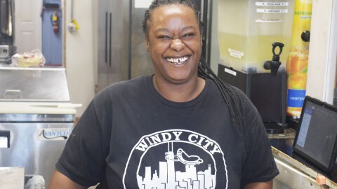 Woman smiling and standing next to a restaurant kitchen and wearing a black shirt with a skyline logo and "Windy City" inscribed on the front.