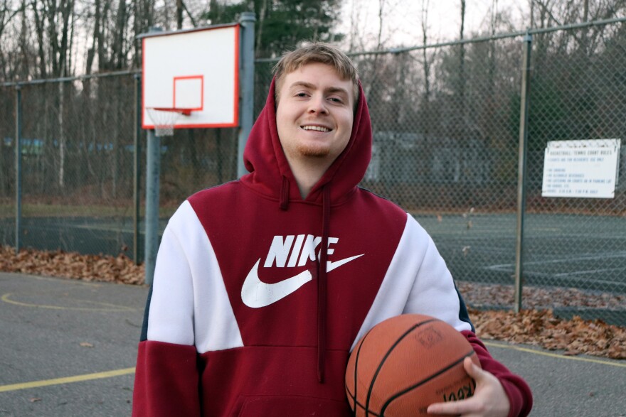 A man poses on a basketball court holding a basketball and smiling