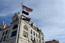 A flag pole stand in front of the Cole County Courthouse. The American flag, the POW flag and the Missouri state flag blow in the wind. The courthouse in the background is gothic architecture, white brick. 