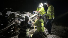 Four men in safety shirts work on a machine in the dark, illuminated with headlamps. 
