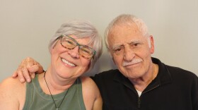 Kay and Arnold Martinez share a sideways hug at their StoryCorps appointment in St. George, Utah.  Kay has short white-gray hair and wears glasses with colorful frames, blue and black on the top tan and black on the bottom.  She smiles with one arm under her father's arm. She is wearing an army green tank top, and a large green pendant hanging from a dark cord around her neck.  Arnold has one hand on Kay's shoulder, and his thumb is holding his index finger.  Arnold has white hair and mustache and wears a black  pullover with a short zipped placket, a left chest pocket with a button, an da piece of paper  slightly extending from the pocket.  Arnold looks toward the camera in a matter-of fact way, his dark eyes expressing love, and his mouth in a straight line.
