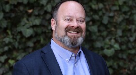 a white man with facial hair wearing a light blue shirt and dark blue suit jack smiles and stands in front of a green ivy background