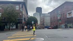 A crossing guard, wearing a reflective vest and hat holds a stop sign as people cross at a crosswalk.