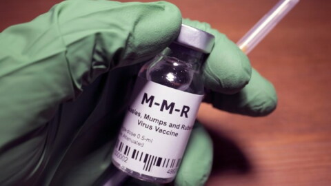 A hand in a green medical glove holds a vial labeled "M-M-R" and a syringe