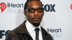 A black man with short hair and wearing a leather black and white tuxedo stares right into the camera with a serious expression on his face.
