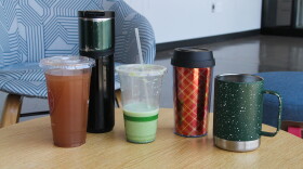 Five coffee cups sit on a table: a clear plastic single-use cup, a tall reusable to-go cup, a half-empty, clear plastic single-use cup, a smaller reusable to-go mug, and a metal mug with a handle. 