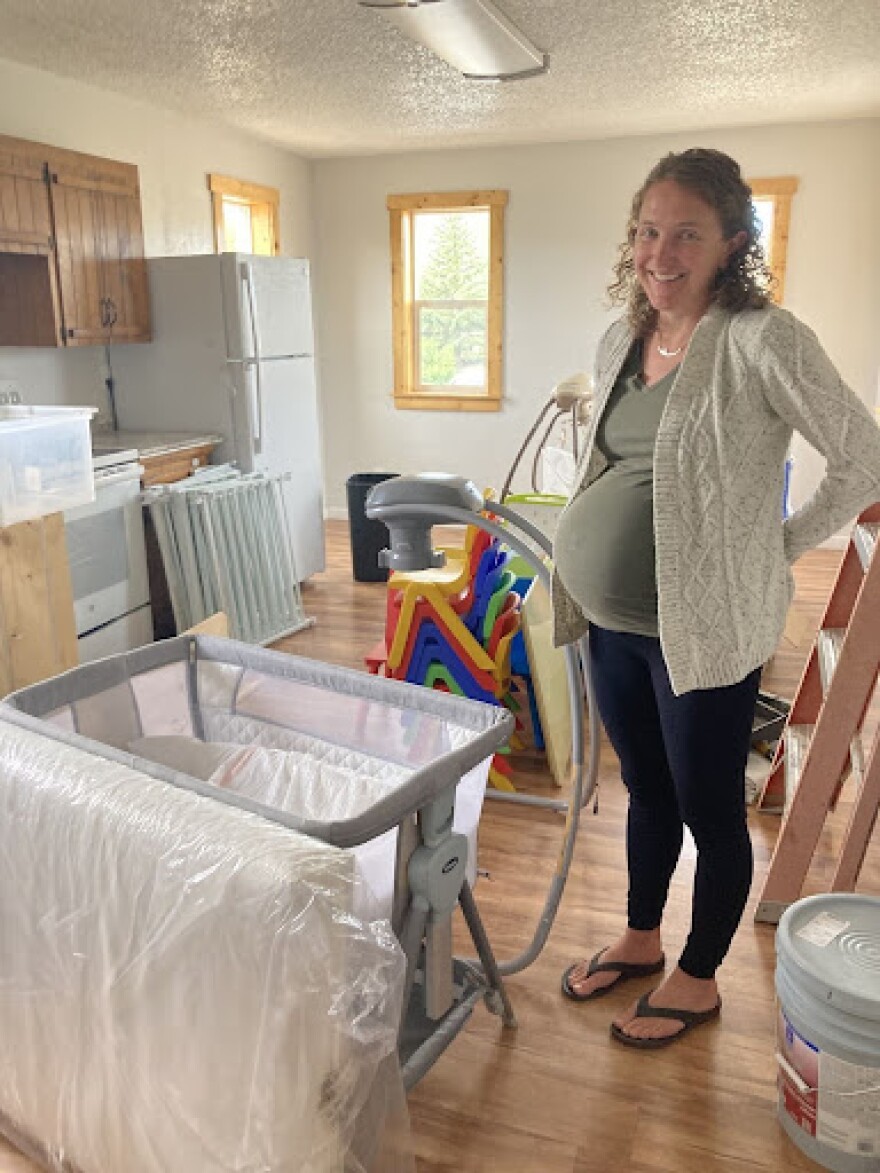 A very pregnant woman stands next to a new crib in a room filled with other new-looking supplies.