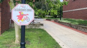 Octagon shaped road sign with the words "Walk your wheels Dismount Zone" affixed to a pole. 