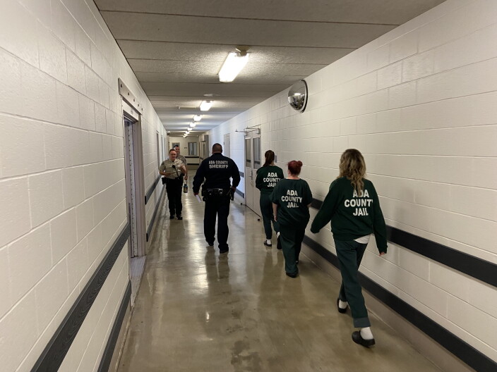 Five people walk down a long hallway with a concrete floor and white cinderblock walls. Four of the people are waring all black with the words "ada county jail" on them. 