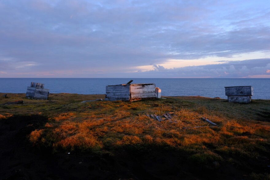 Ice cellars, constructed of wood boards, rest on a grassy field with the ocean looming in the background.