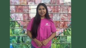 A young woman with long dark hair wearing a pink t-shirt with the Boys and Girls Club logo stands in front of a tiled wall with a colorful mosaic. 