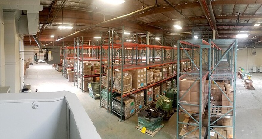 The interior of Spearpoint Logistics. There are rows of metal shelving units placed as aisles in a warehouse. Most shelving units include stacked cardboard boxes. There are also plastic-wrapped pallets placed between many of the aisles.