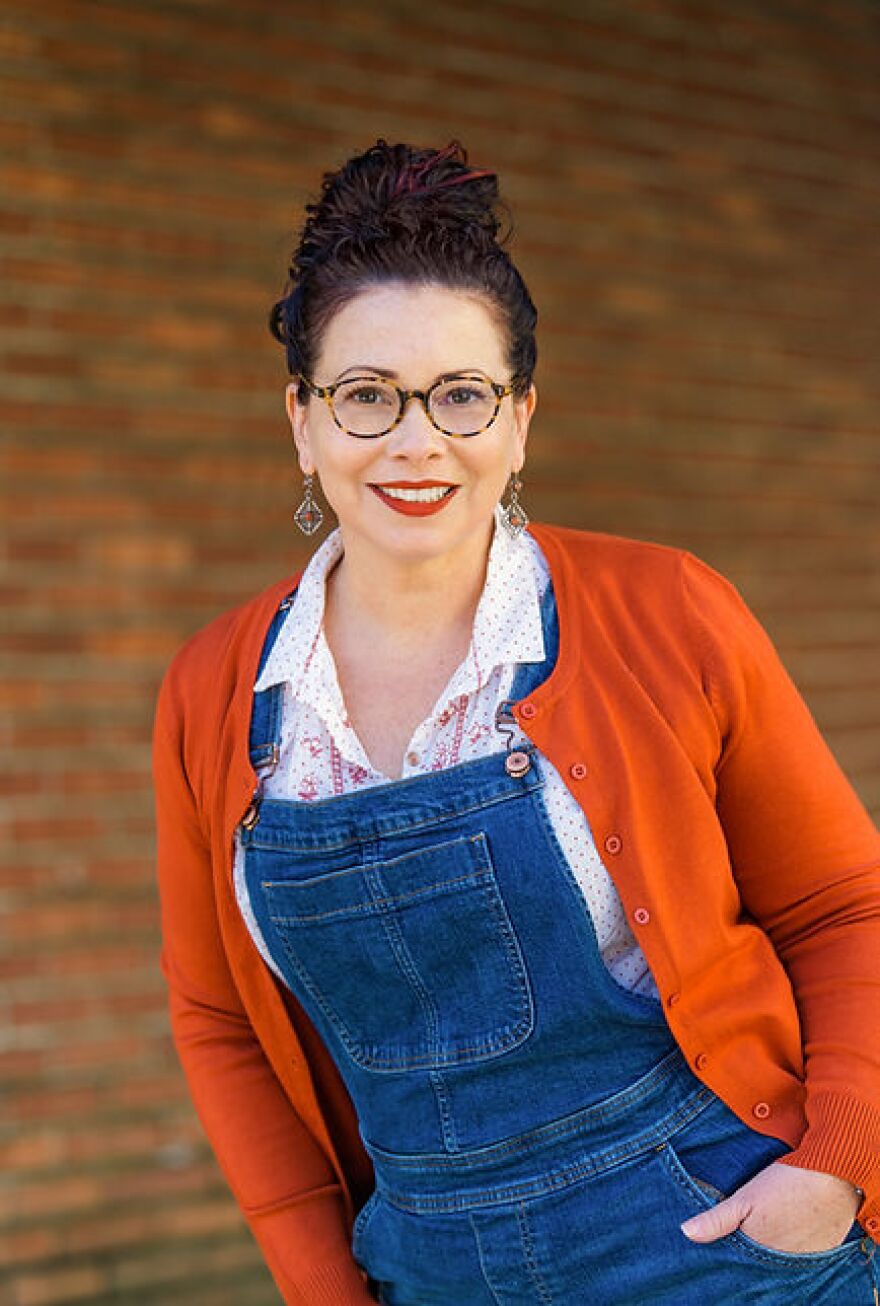 Donna Barba Higuera wears denim overalls, a white blouse and a red-orange sweater. Her hair is in a bun atop her head.